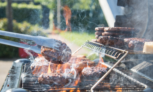 A two-tiered propane grill with burgers cooking on the lower grate and others staying warm on the top shelf. Tongs are flipping one burger, while flames rise from the grill grates.